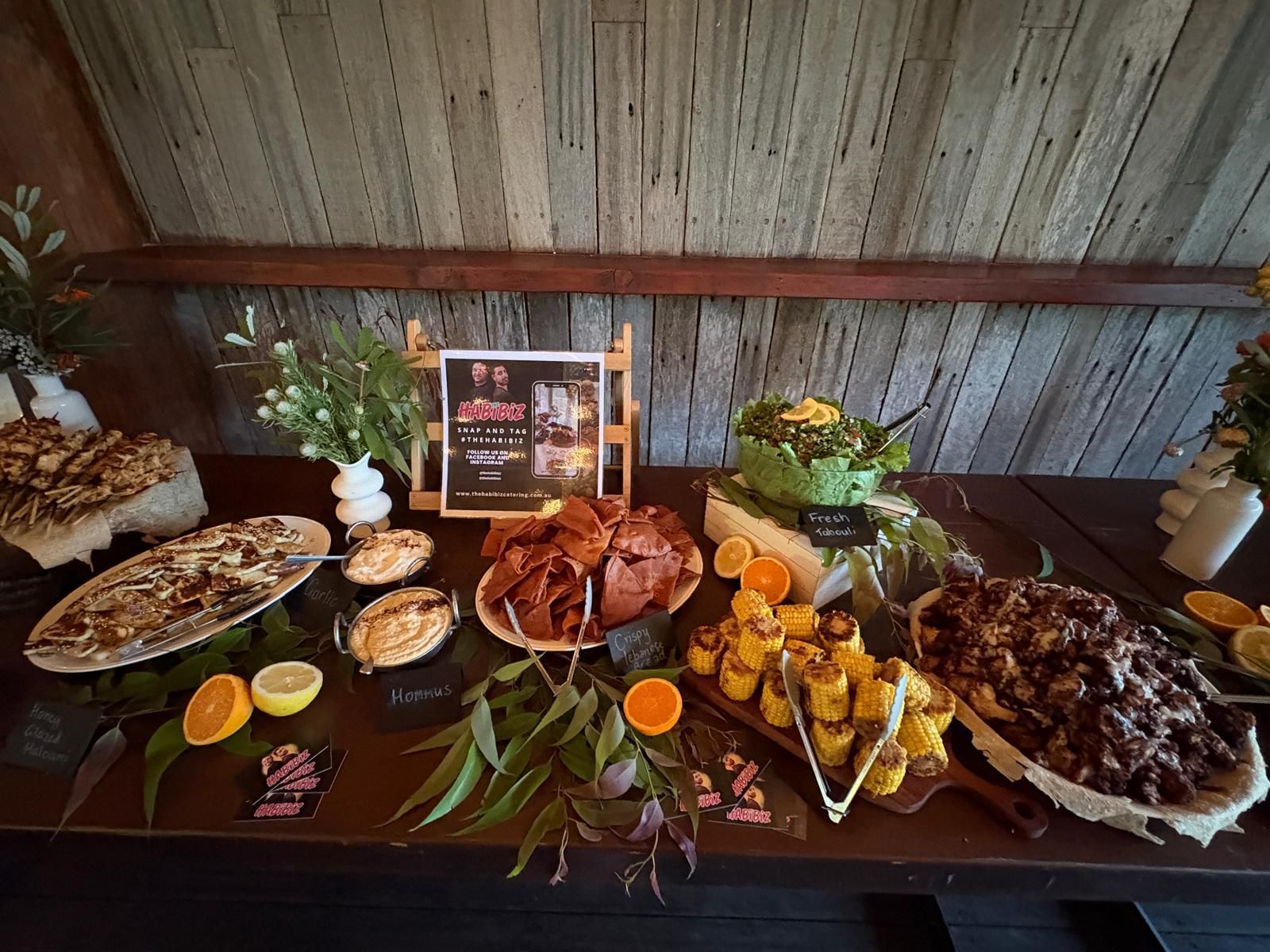 A table laden with appetizers, including dips, pastries, and orange slices, is set against a wooden backdrop. — The Habibiz Catering In Wingham, NSW