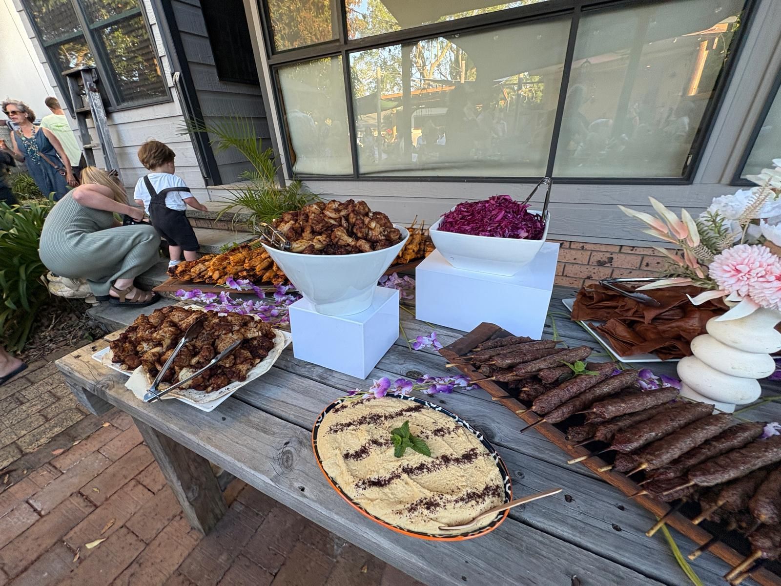 Outdoor buffet table with various foods: skewers, fried items, hummus, and salad. People in background. — The Habibiz Catering In Wingham, NSW