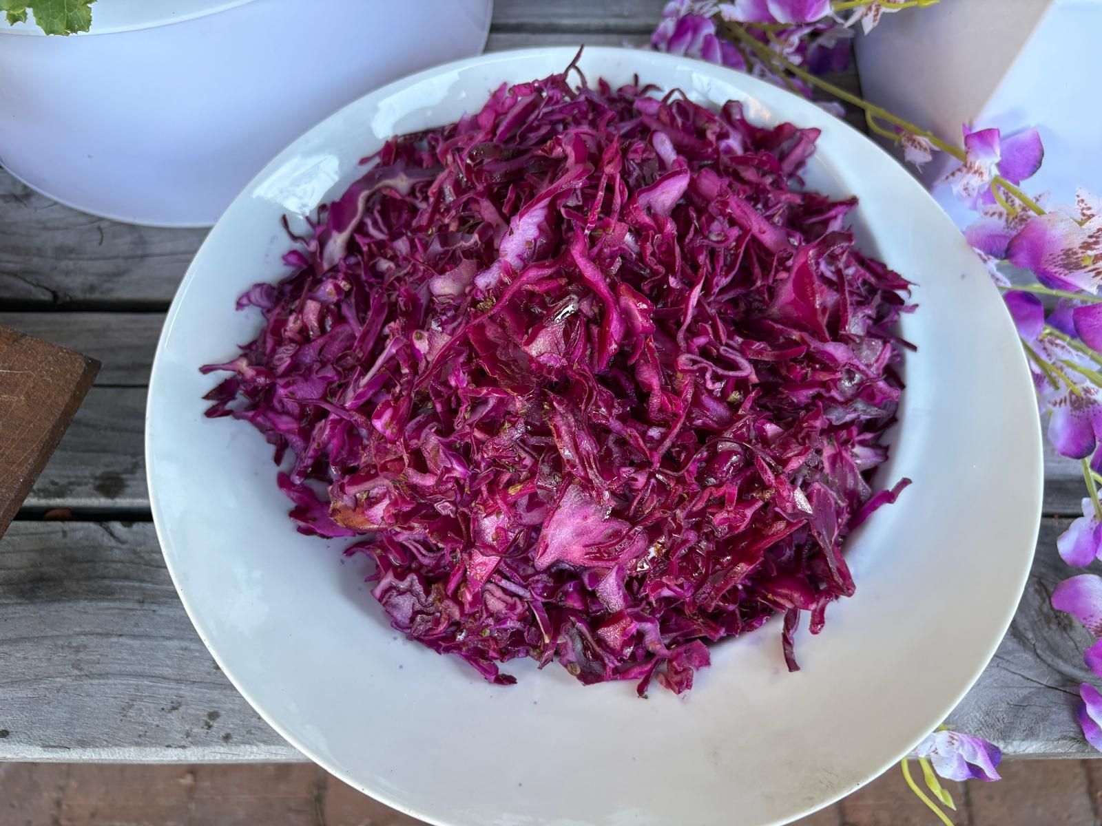 Shredded red cabbage salad in a white bowl, on a wooden surface, with purple flowers. — The Habibiz Catering In Wingham, NSW