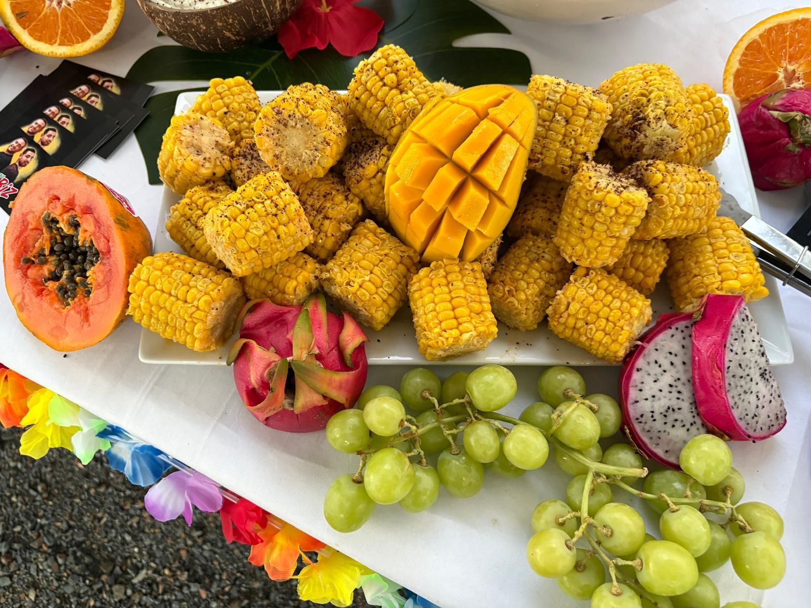 A platter of grilled corn, a mango, papaya, dragon fruit, and grapes, arranged on a decorated table. — The Habibiz Catering In Wingham, NSW