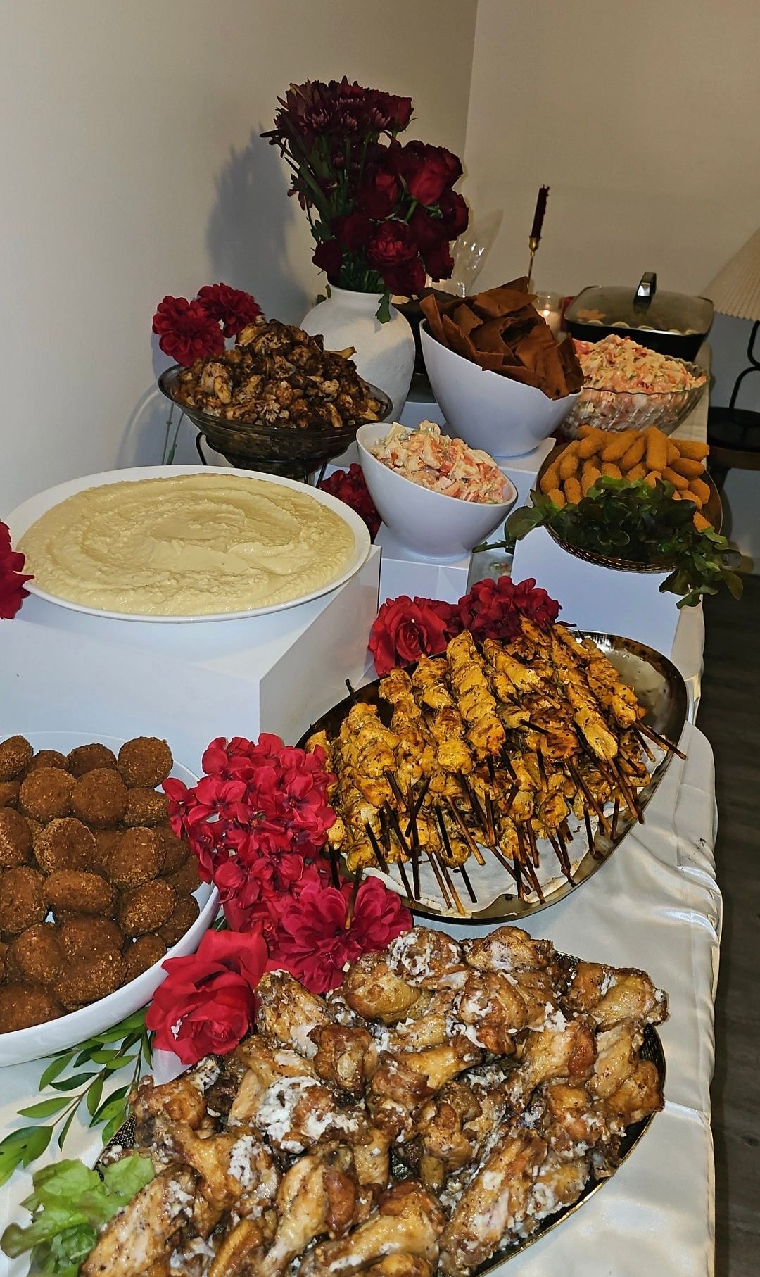 A buffet table with various food dishes, including skewers and chicken wings, decorated with red flowers. - The Habibiz Catering in Wingham NSW