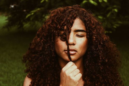 Person with curly hair holding a strand over their face against a dark green background