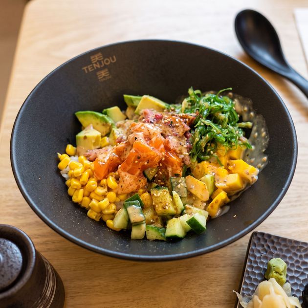 Black bowl of mixed salad grains, corn, avocado, salmon, and greens on a wooden table with a spoon.