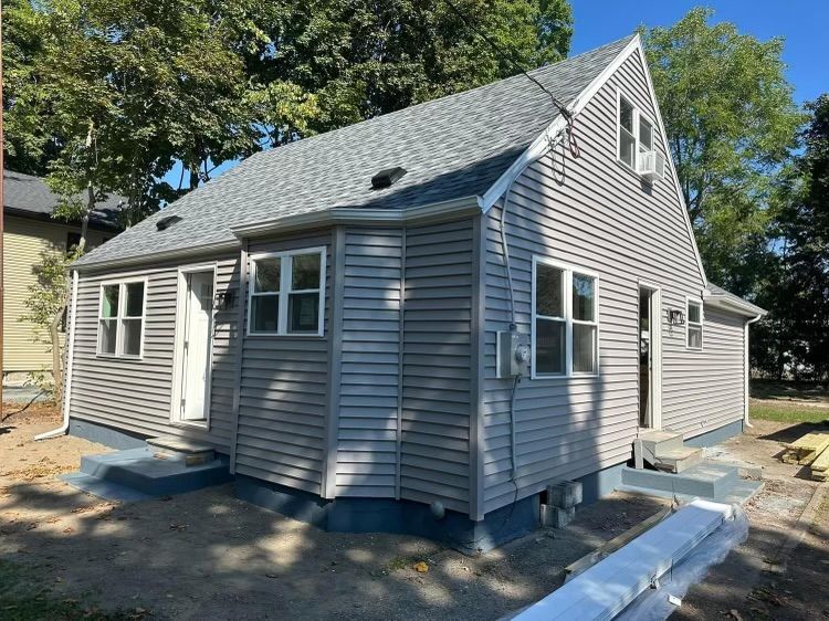 Small gray house with white trim and a steep roof, shown from the front in a sunny yard