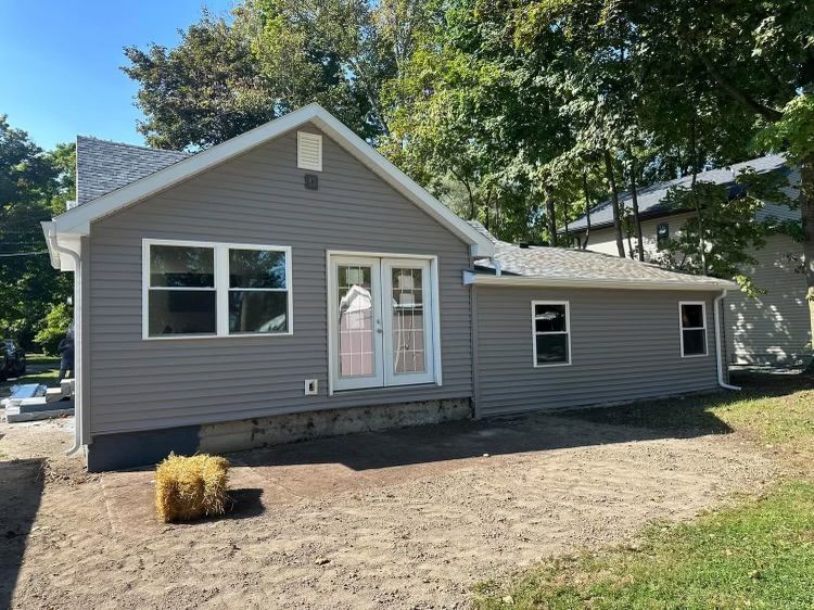 Gray house with white trim, front door, and side addition on a bare yard under trees
