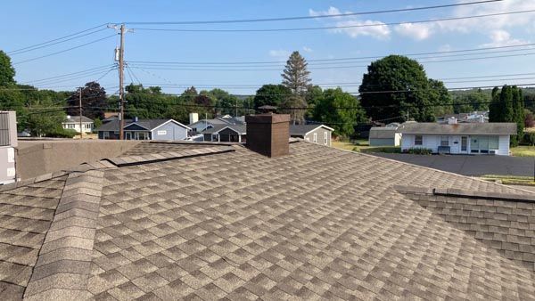 Rooftop with tan shingles and a chimney, overlooking suburban houses and trees under a partly cloudy sky