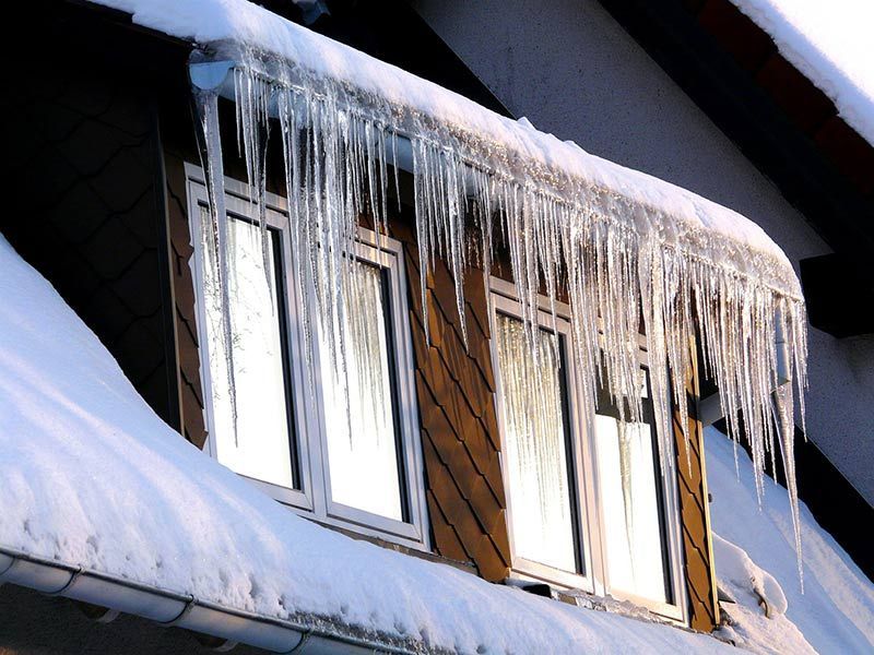 Snow-covered house roof with long icicles hanging from the eaves and windows below.