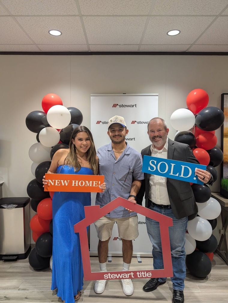 Three people pose at a real estate closing with SOLD sign, New Home sign, and house frame prop.