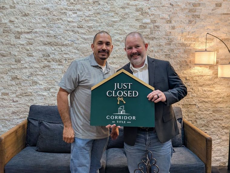 Two men hold a “Just Closed” sign in front of a stone wall and couch.