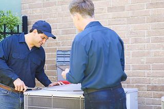 Two technicians in blue uniforms inspect an outdoor air conditioner unit beside a brick wall.