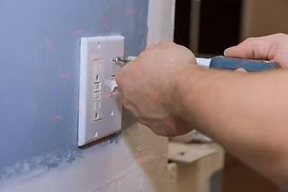 Hand tightening a white electrical wall outlet plate with a blue screwdriver