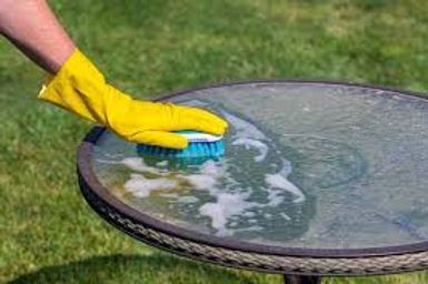 Gloved hand scrubbing a glass patio table with a blue sponge outdoors.