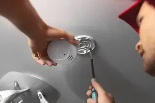 Hand using a screwdriver to remove a round smoke detector from a ceiling mount