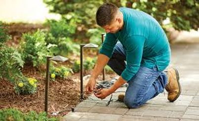 Person kneeling on a garden path, adjusting stone pavers beside green plants and lantern lights