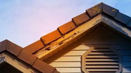 Sunlit house roof with brown shingles and a round attic vent under a blue sky