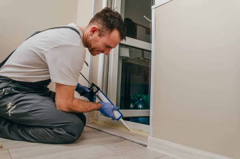 Man in gray shirt sealing the baseboard with a caulking gun beside a wall and window.