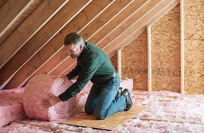 Man kneeling in an attic, placing pink fiberglass insulation between rafters