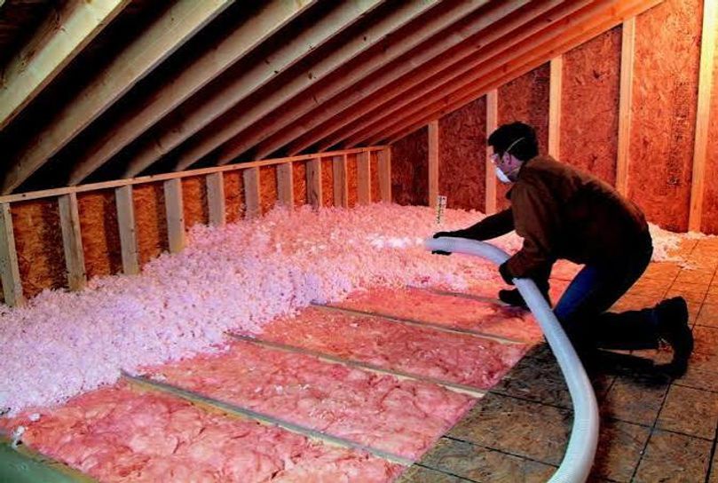 Person kneeling in an attic, blowing loose insulation into floor joists with a hose