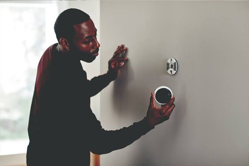 Person installing a wall-mounted smoke detector on a blank wall with a drill in hand