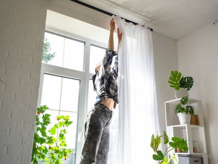 Person reaching up to hang white curtains by a bright window in a plant-filled room