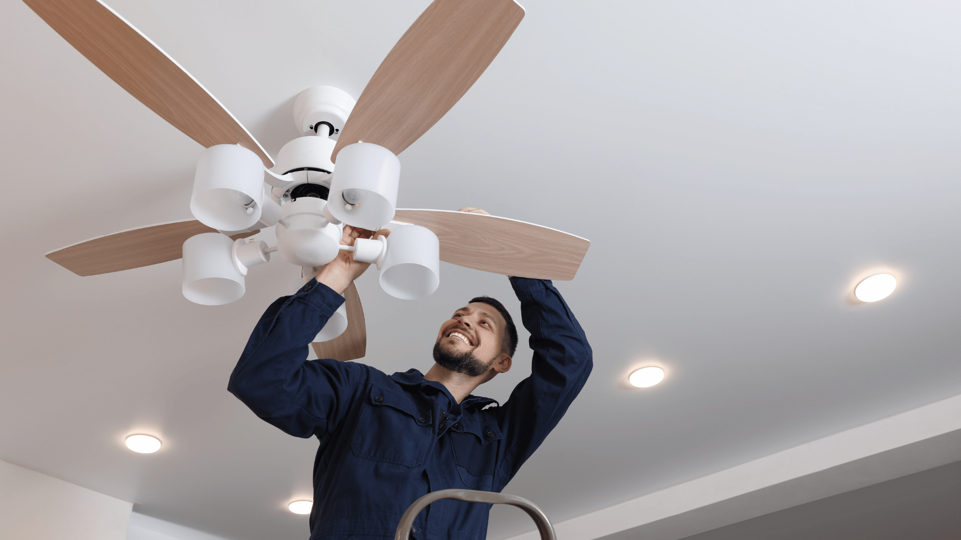 Man installing a white ceiling fan with wood blades on a ceiling indoors.