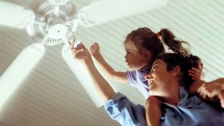 Two people adjusting a ceiling fan in a bright room, looking upward