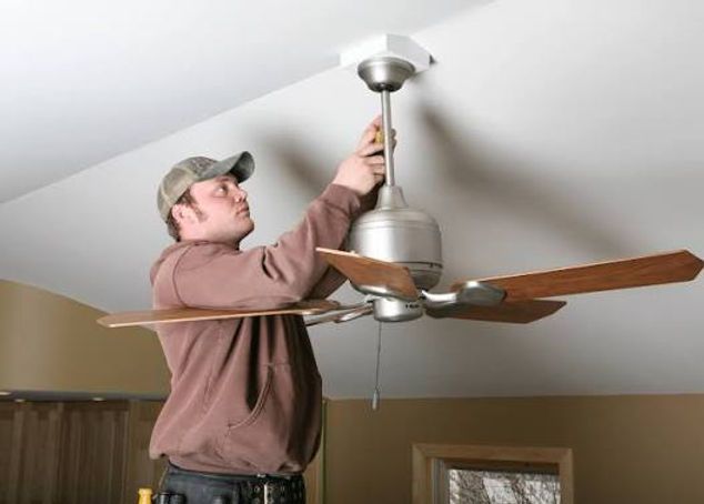 Man installing a ceiling fan with wooden blades in a room with a sloped ceiling