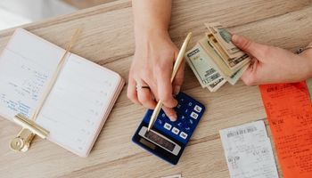 Hands counting cash beside a smartphone and calculator on a desk with notebooks and receipts