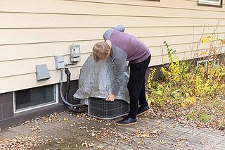 Person bending over an outdoor HVAC unit beside a house, inspecting it near a wall and shrubs.