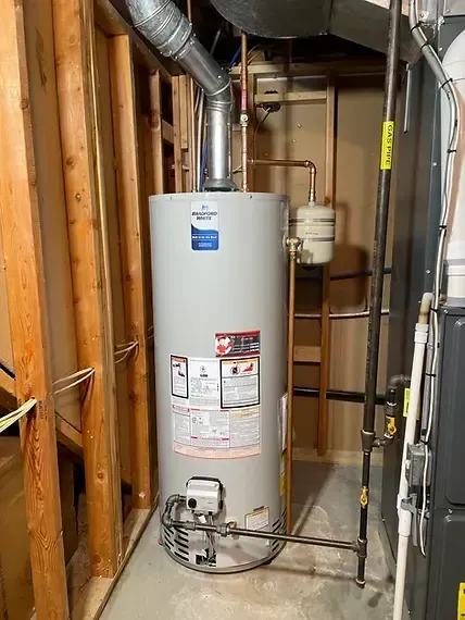 Water heater in a utility closet with exposed pipes and ductwork, beside a furnace and unfinished wooden framing.