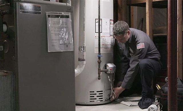 Technician kneeling beside a water heater, inspecting a pipe connection in a utility room