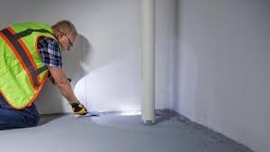Worker in a high-visibility vest applying sealant along the floor-wall seam in a room.