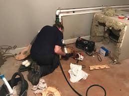 Person kneeling on floor beside a fireplace, working with tools and cables in a room.