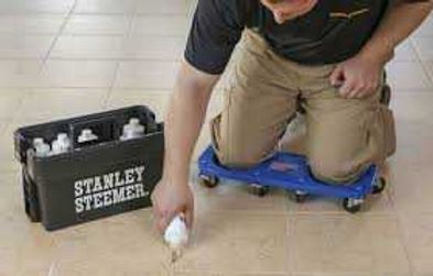 Person kneeling on a blue kneeling scooter, reaching for a white bottle beside a Stanley Steemer box.