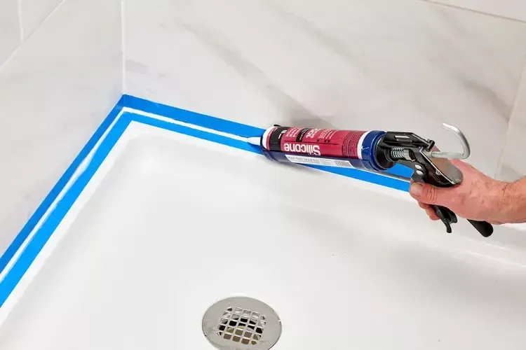 Hand applying caulk along a bathtub seam masked with blue painter’s tape