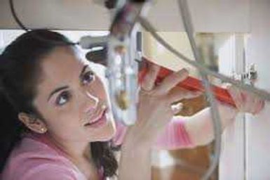 Woman in a pink shirt adjusting pipes under a sink, looking upward with a slight smile.