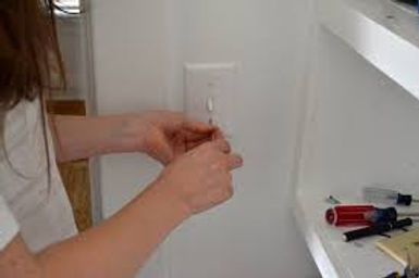 Hands plugging a white cord into a wall outlet beside a windowsill with small tools nearby