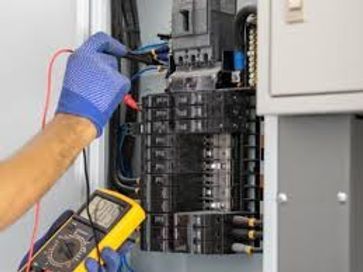 Gloved electrician testing wires in an open electrical panel with a yellow multimeter