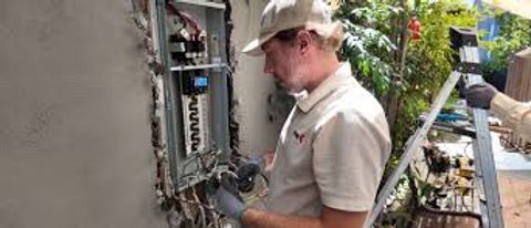 Technician working on an electrical panel outdoors beside a house, wearing a cap and beige shirt