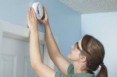 Woman reaching up to adjust a white smoke detector on a wall near the ceiling