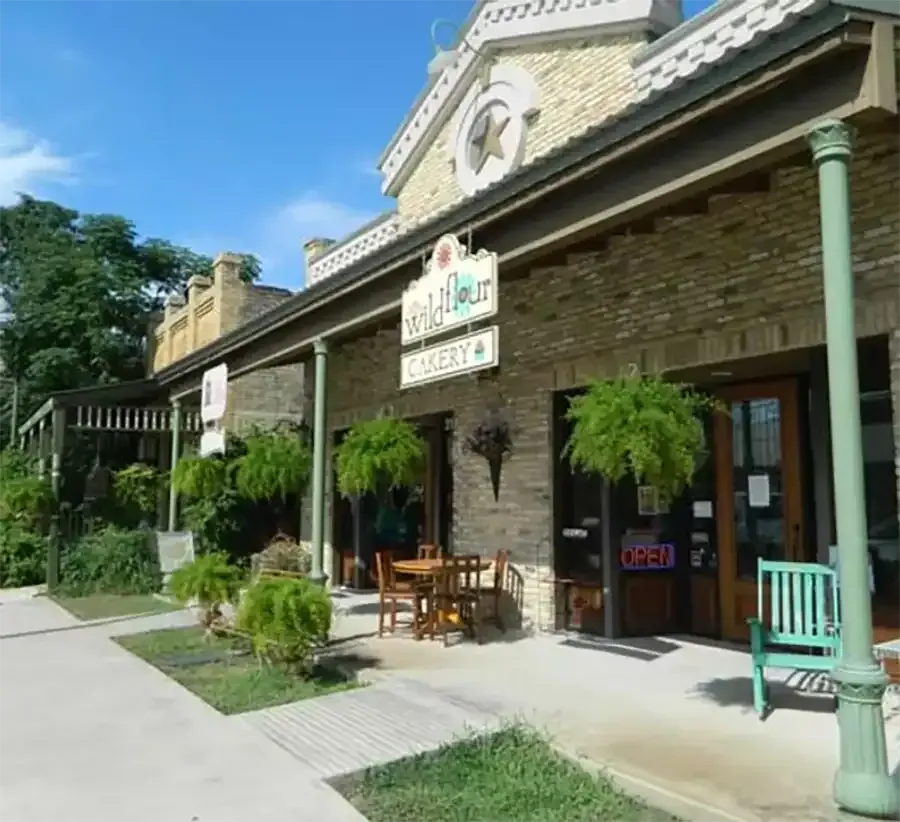 Historic brick storefront with green trim, hanging ferns, and outdoor seating along a sunny sidewalk.