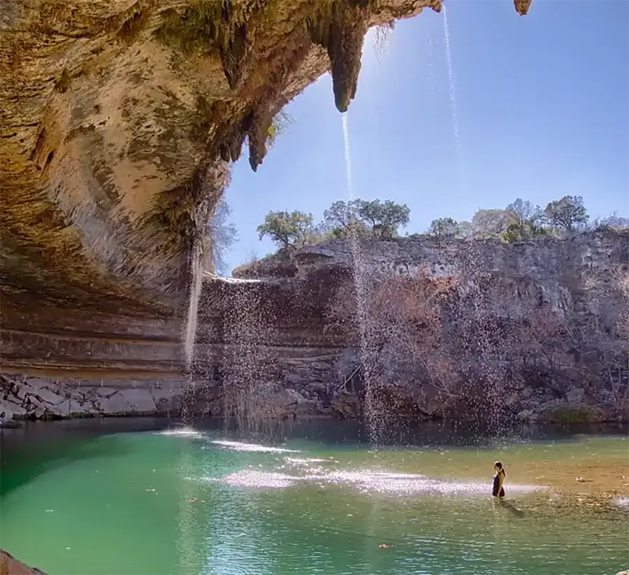 Waterfall spilling into a turquoise pool inside a rocky cave, with a person wading near the shore