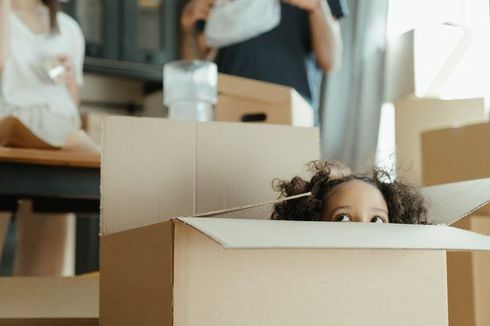 Child peeking out of a cardboard box in a room with moving boxes and people in the background