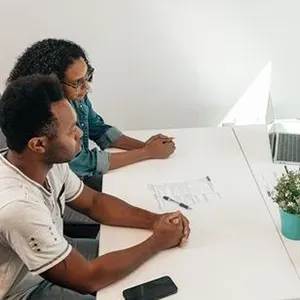 Three people in a bright office meeting around a white table with papers and a laptop, discussing a sketch.