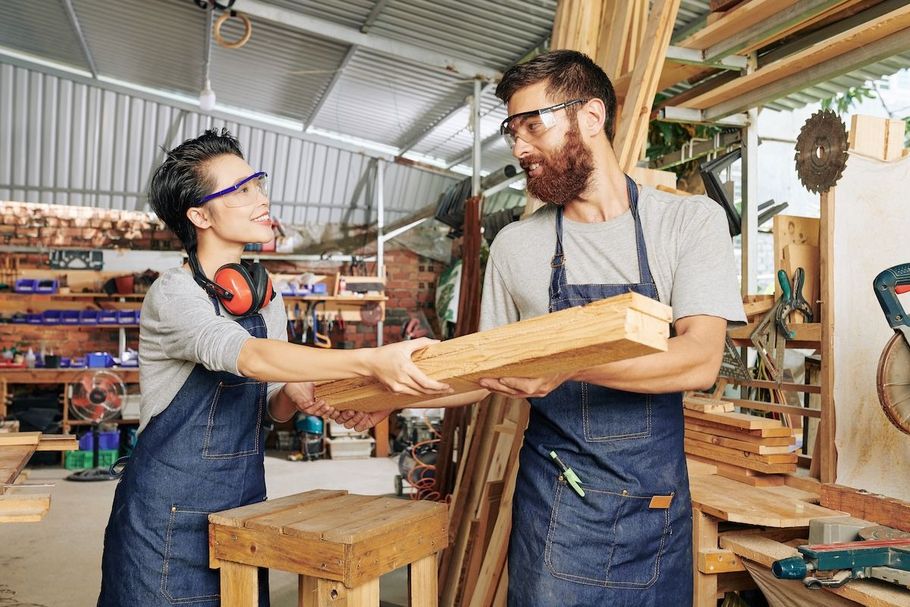 Two woodworkers passing a wooden board in a workshop, wearing aprons and safety glasses.