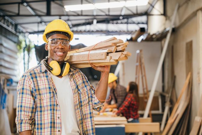 Worker in a yellow hard hat carrying lumber in a workshop, with wood supplies stacked nearby