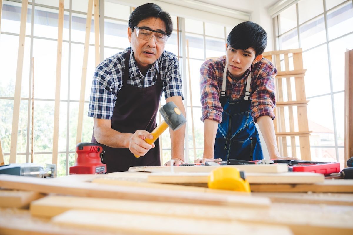 Two people in aprons hammering wood in a bright workshop with tools on a workbench
