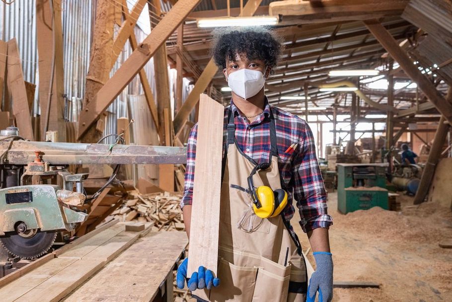 Masked worker in a wooden workshop, holding a long plank and wearing overalls, gloves, and a tape measure.
