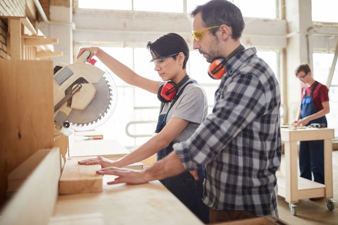 Two people working with a circular saw in a bright woodshop, cutting wood on a workbench
