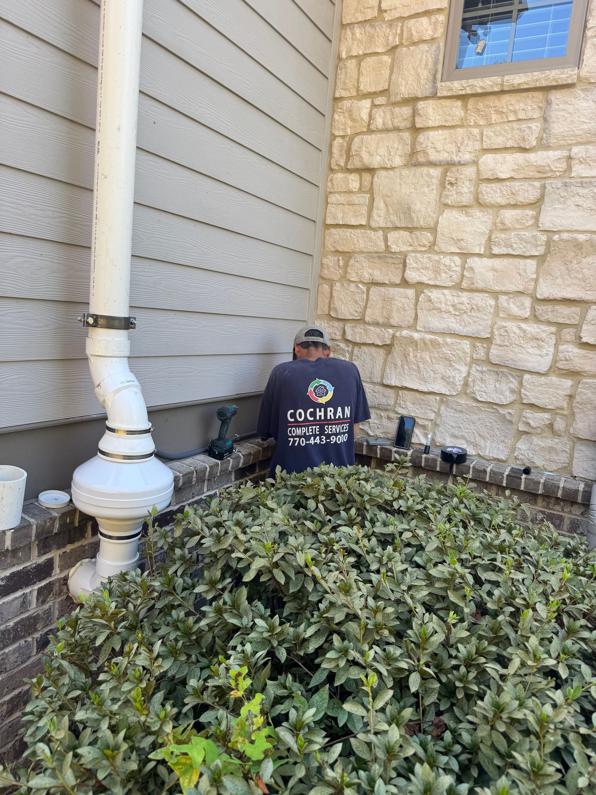 Person in a dark shirt working near a house's exterior. White pipe, stone and light-colored siding. Bush in the foreground.